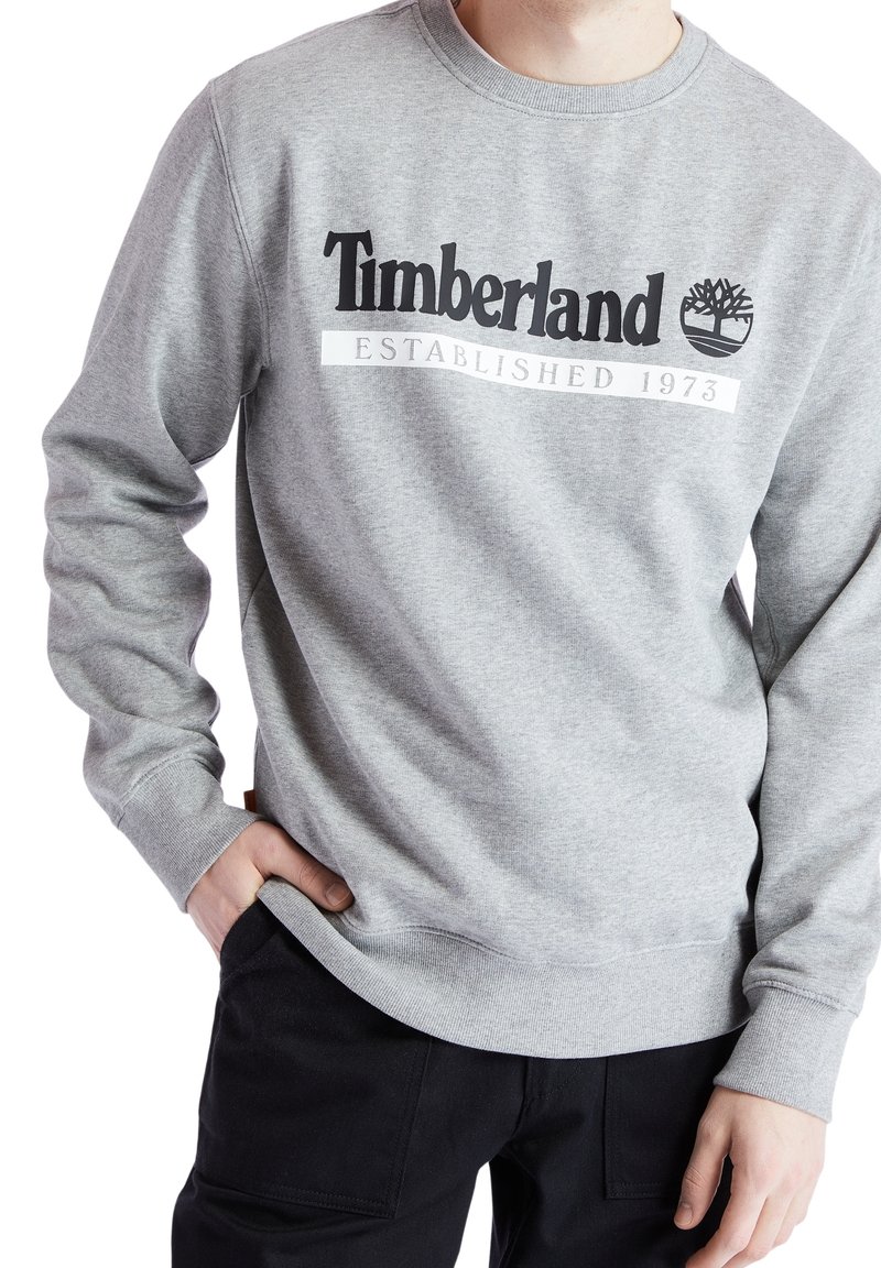 Personne portant un sweat Timberland gris avec logo et texte "Established 1973", associé à un pantalon noir, main dans la poche.