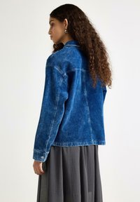 Modrá džínová bunda s vybledlým efektem, manžetami na knoflíky a klasickým límcem, viděná zezadu. Dlouhé kudrnaté vlasy padají přes rameno.