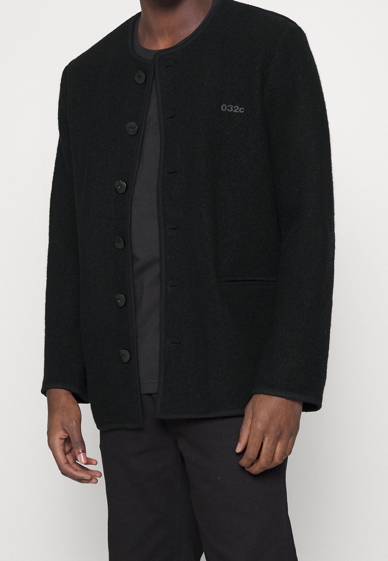Veste en laine noire avec un col rond, fermeture à boutons sur le devant et manches longues. Présente un logo discret et un tissu doux et texturé.