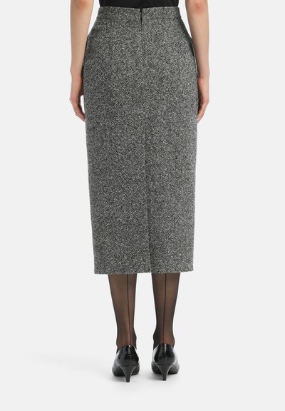 Egy nő magas derékkal rendelkező, szürke tweed midi szoknyát visel, fekete varrott harisnyával és fekete, alacsony sarkú cipővel, háttal a kamerának.