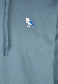 Felpa leggera blu con cappuccio, caratterizzata da un motivo ricamato di un uccello bianco e blu sul petto. Realizzata in un morbido tessuto testurizzato con cappuccio regolabile tramite cordino.
