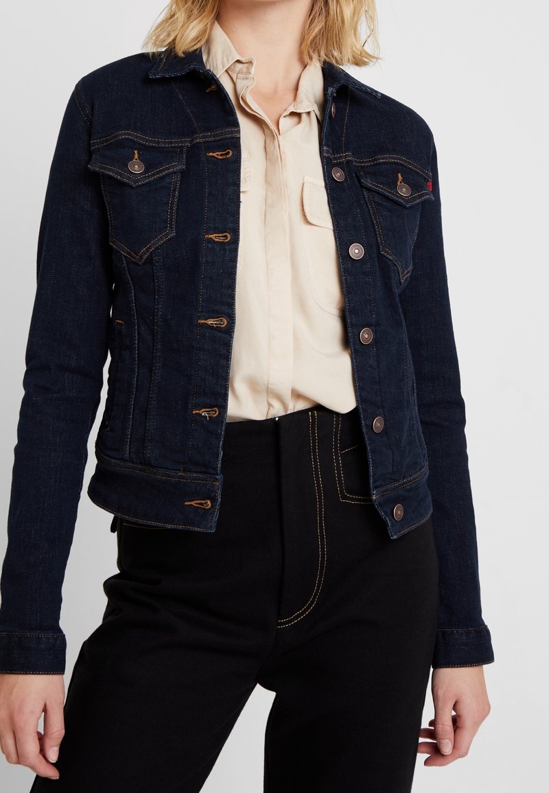 Veste en denim foncé avec deux poches poitrine, boutons métalliques et coutures contrastées. Portée par-dessus une chemise beige, associée à un pantalon noir.