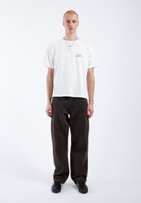 T-shirt en coton blanc avec une petite signature noire sur le côté gauche de la poitrine, associé à un pantalon ample foncé et des chaussures noires.