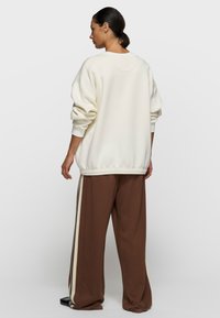 Sudadera crema con ajuste extragrande, puños y dobladillo acanalados. Pantalones marrones con rayas laterales crema. Tejido suave, diseño casual, silueta relajada.