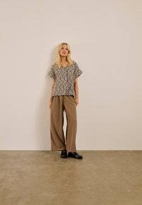 Femme debout contre un mur beige, portant un chemisier à manches courtes à motifs, un pantalon large marron et des mocassins noirs sur un sol en béton.