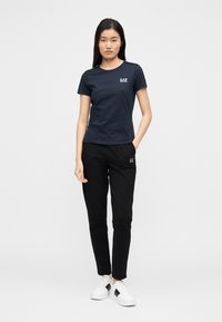 Marineblauw T-shirt met een ronde hals en logo, gecombineerd met een zwarte broek en witte sneakers, voorzien van een elastische tailleband en zakken.