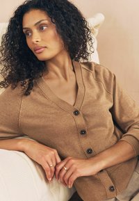 Cardigan en tricot marron avec un col en V, fermeture à boutons et manches courtes. Texture lisse avec des accents côtelés au niveau de l'ourlet et des poignets.