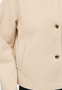 Manteau en laine beige à l’avant avec deux boutons marron et une poche latérale fendue, manche partiellement visible sur le côté gauche.