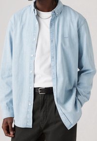 Man in lichtblauw denim overhemd met knopen over een wit T-shirt, zwarte broek, zwarte riem en zilveren ketting, staat met één hand in zijn zak.