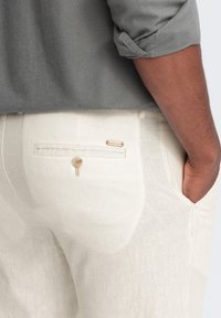 Pantalons en lin beige avec poches et fermeture à bouton. La vue arrière montre une texture subtile et une petite étiquette de marque près de la poche droite.
