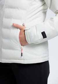 Biała kurtka puchowa o pikowanej strukturze, boczna kieszeń z różowym zamkiem błyskawicznym i czarną łatą z logo na rękawie. Posiada gładkie wykończenie.