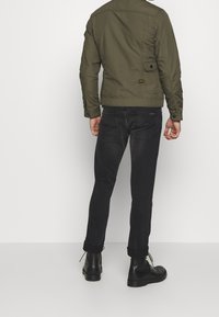 Man in een olijfgroene jas, zwarte opgerolde slim fit jeans en zwarte leren boots, staand tegen een effen witte achtergrond.