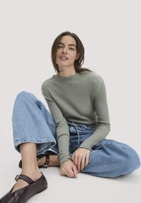 Khakifarbenes, geripptes Langarmoberteil, kombiniert mit hellblauen, weit geschnittenen Jeans. Die Schuhe sind dunkelbraun, flach und mit einem Knöchelriemen. Sitzpose.