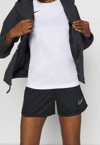Sporty outfit featuring a white short-sleeve shirt, black shorts with a green Nike swoosh, and a dark jacket with zipper.