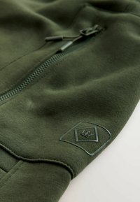 Olivgrüner Sweatshirt mit Reißverschluss, gerippten Bündchen und einem strukturierten Logo-Patch am Ärmel. Weicher, glatter Stoff.