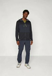 Tume sinine kapuutsiga jakki, kontrastse heleda halli rinnaosaga, lukkuga taskud, koos tume siniste lahtiste pükste ja hallide tossudega.