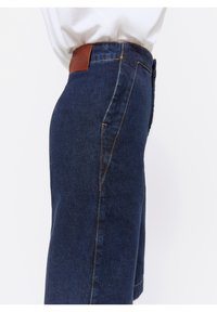 Lähikuva tummansinisestä farkkuhameesta, jossa on keltaisia saumoja ja ruskea nahkavalmisteinen bränditunnus vyötäröllä, käytettynä valkoisen paidan kanssa.