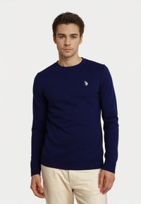 Joven vestido con un suéter de manga larga azul marino con un pequeño emblema en el pecho y pantalones de color claro, de pie frente a un fondo blanco.
