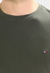 Mężczyzna ubrany w oliwkową koszulkę z okrągłym dekoltem, z małym prostokątnym, wyhaftowanym logo w kolorach czerwonym, białym i niebieskim po prawej stronie klatki piersiowej.