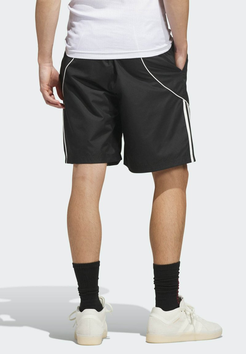 Personne portant un short de sport noir avec un liseré blanc, des baskets blanches, des chaussettes noires et un débardeur blanc, debout les mains dans les poches.