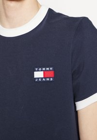 Marineblaue Baumwoll-T-Shirt mit weißem Kragen, ausgestattet mit einem rechteckigen gestickten Logo-Patch in Rot, Weiß und Blau, das "TOMMY JEANS" liest.