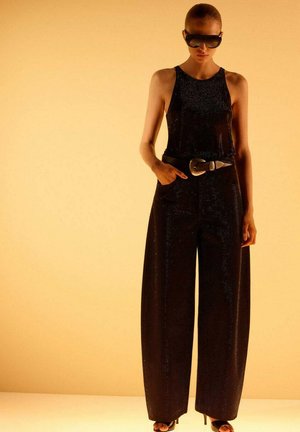 Woman wearing black sleeveless jumpsuit with wide legs, large belt buckle, sunglasses, and high-heeled sandals against a yellow background.