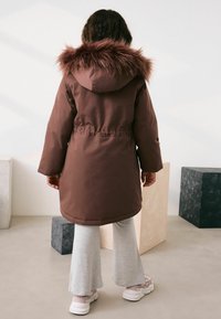 Brun parka med en faux fur hætte, har en justerbar talje og lange ærmer, båret over lysegrå flared bukser og sneakers.