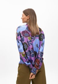 Blusa di satin viola con motivi floreali in blu, verde e rosso scuro; maniche lunghe con polsini a bottoni, vestibilità rilassata, texture liscia.