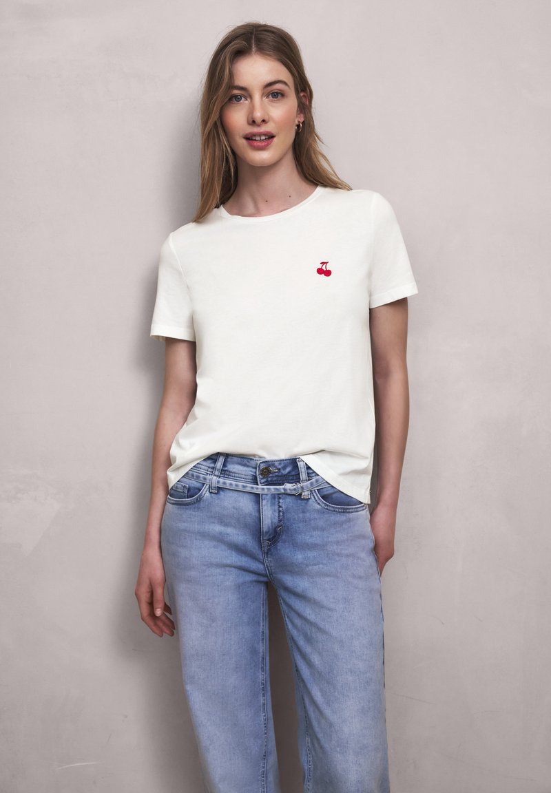 Kobieta ubrana w białą koszulkę z małym czerwonym wzorem wiśni oraz jasnoniebieskie dżinsy, stojąca na tle jednolitej jasnej ściany.