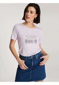 Liliowa koszulka z krótkim rękawem i okrągłym dekoltem, z błyszczącym graficznym napisem "Cross Jeans" oraz wzorem frędzli. Zestawiona ze spódnicą dżinsową.