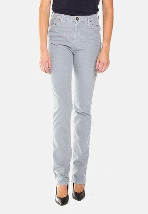 Vrouw die lichtgrijze slim-fit broek en zwarte puntige hakken draagt, staand tegen een effen lichte achtergrond.