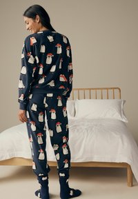 Marineblaue Schlafanzüge mit Pinguin-Muster in roten Hüten, bestehend aus langen Ärmeln und Hosen. Weicher Stoff, kombiniert mit dunklen, kuscheligen Socken. Bett im Hintergrund.