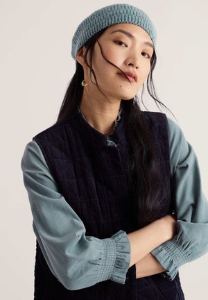 Jeune femme aux longs cheveux foncés portant un bonnet en maille bleu, un chemisier turquoise avec des manches froncées, et un gilet matelassé noir, bras croisés.