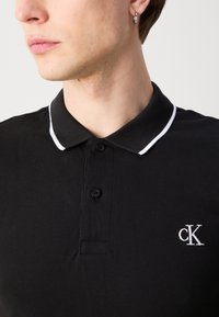 Mladý člověk nosí černé polo tričko s bílým lemováním na límci a malým bílým logem "cK" na hrudi, také nosí stříbrnou kruhovou náušnici.