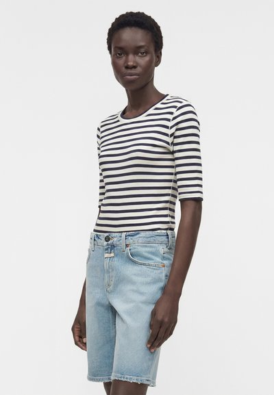 Γυναίκα που φοράει μπλε ναυτικό και λευκό ριγέ μπλουζάκι με μισά μανίκια και ανοιχτό μπλε τζιν σορτς μέχρι το γόνατο, στέκεται μπροστά σε ένα απλό λευκό φόντο.