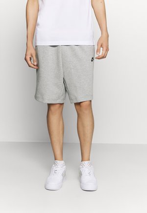 Homme portant un short Nike gris clair, un t-shirt blanc et des baskets blanches, debout devant un fond clair uni.