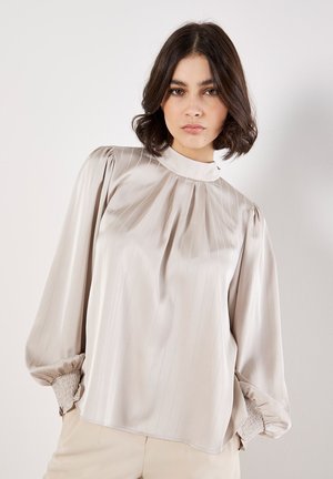 Blusa beige chiaro realizzata in tessuto liscio con righe verticali, collo alto e maniche a sbuffo con polsi arricciati.
