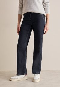 Pantalones de denim azul oscuro con corte recto, detalles de costura sutiles, combinados con zapatillas deportivas blancas. La tela tiene una textura suave.