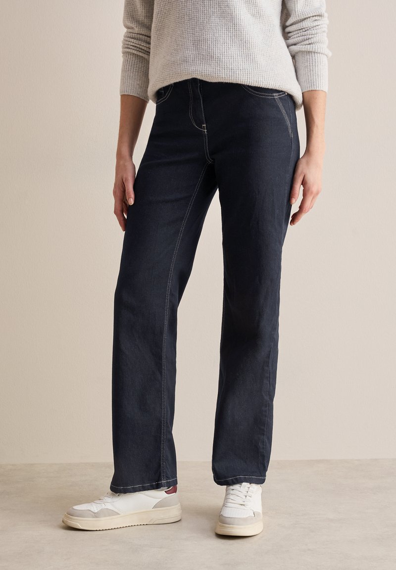 Pantalones de denim azul oscuro con corte recto, detalles de costura sutiles, combinados con zapatillas deportivas blancas. La tela tiene una textura suave.