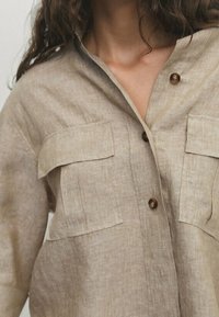 Beige linned skjorte med krave, to brystlommer og træknapper. Stoffet har en tekstureret, let krøllet udseende.