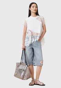 Parte superior blanca con detalles de encaje y malla, pantalones cortos de mezclilla, bolso metálico plateado y sandalias planas con tiras. Contrastes suaves y texturizados.