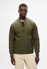 Giubbotto bomber trapuntato verde oliva con chiusura frontale a zip, polsini a costine e due tasche laterali. Il modello indossa un top beige sotto.