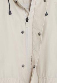 Giacca beige frontale con zip bianca centrale, cordini neri con estremità in plastica e bottoni a pressione in metallo lungo la patta.