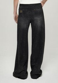 Fekete bővülő szárú nadrág szegecselt díszítéssel. Simább textúrájú, jól látható fémes részletekkel és egy barna címkével a hátsó zsebén.