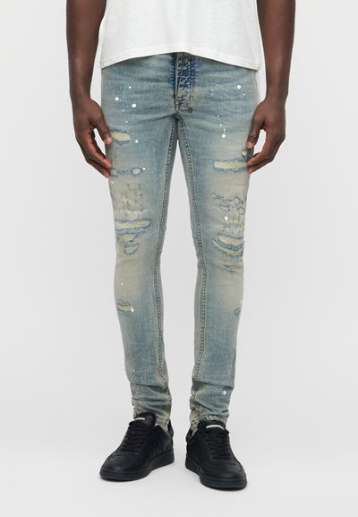 Jean skinny déchiré en denim bleu clair avec des détails de taches de peinture et plusieurs déchirures. Associé à des baskets noires.