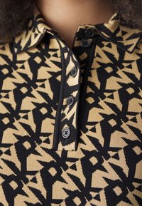 Collet et patte de boutonnage d'un pull noir et beige à motif géométrique porté par une personne aux cheveux bouclés.