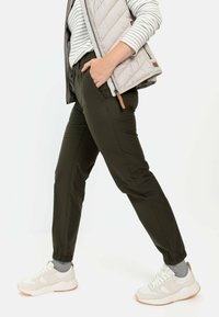 Dunkelgrüne, schmal geschnittene Hose mit elastischen Bündchen, kombiniert mit weißen Sneakern und einer hellgrauen Steppweste über einem gestreiften Hemd.