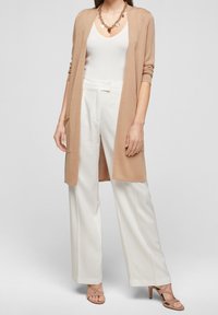 Beige lång kofta med öppen front, sidofickor, bärs över en vit linne och kombineras med vida vita byxor och sandaler med remmar i beige.