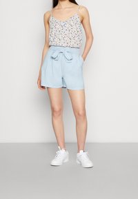Hedvábný top s květinovým vzorem a tenkými ramínky, spárovaný s měkkými modrými šortkami s vysokým pasem, které mají přední mašličku a kapsy. Bílé tenisky.