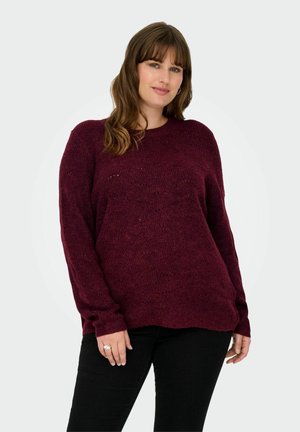 Donna con capelli castani di lunghezza spalle, che indossa un maglione a maglia bordeaux e pantaloni neri, in piedi con un lieve sorriso su uno sfondo semplice.